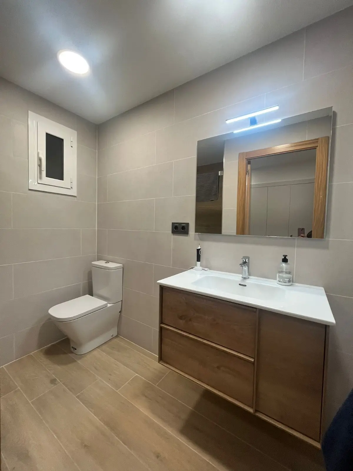Baño con mueble suspendido de madera, inodoro compacto y porcelánico gris claro