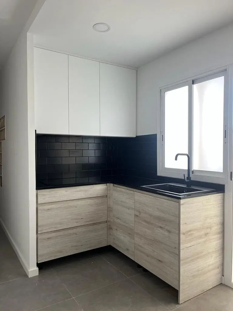 Cocina en L con muebles de madera clara, altos blancos y azulejo metro negro