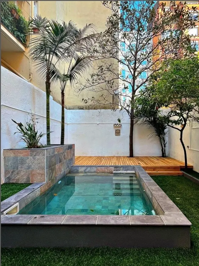 Piscina de obra revestida de piedra con tarima, césped y palmeras en patio urbano